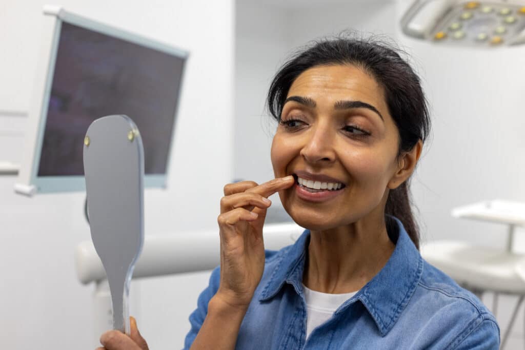 Woman examining her smile in a dental office while considering dental implants and dentures for tooth replacement.